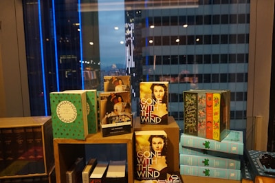 A collection of books is arranged on a display shelf in front of a window with a cityscape view. Dominant books include 'Gone With the Wind' and various colorful, decorative covers. The city buildings outside are lit up with blue and white lights, contributing to the ambient nighttime atmosphere.
