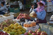 A market scene featuring a vendor sitting behind an assortment of colorful fruits, such as mangoes, dragon fruits, grapes, and others. The vendor is wearing a sun-protective hat and face covering while holding a plastic bag of fruit. Various boxes and crates are filled with produce surrounding the vendor. Another person is visible in the background, sitting and waiting.