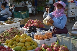 A market scene featuring a vendor sitting behind an assortment of colorful fruits, such as mangoes, dragon fruits, grapes, and others. The vendor is wearing a sun-protective hat and face covering while holding a plastic bag of fruit. Various boxes and crates are filled with produce surrounding the vendor. Another person is visible in the background, sitting and waiting.