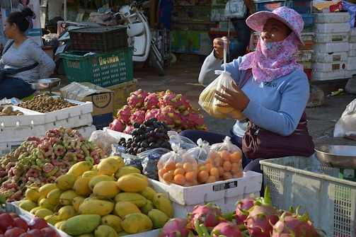 A market scene featuring a vendor sitting behind an assortment of colorful fruits, such as mangoes, dragon fruits, grapes, and others. The vendor is wearing a sun-protective hat and face covering while holding a plastic bag of fruit. Various boxes and crates are filled with produce surrounding the vendor. Another person is visible in the background, sitting and waiting.