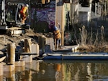 Two construction workers are involved in infrastructure development beside a body of water. One worker is positioned on the edge of the embankment, wearing a helmet and an orange safety vest, preparing machinery. The other worker is on a floating platform, guiding or holding a large metal sheet beside tall reeds. Graffiti decorates the brick wall in the background, adding an urban element to the environment.