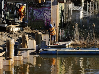 Two construction workers are involved in infrastructure development beside a body of water. One worker is positioned on the edge of the embankment, wearing a helmet and an orange safety vest, preparing machinery. The other worker is on a floating platform, guiding or holding a large metal sheet beside tall reeds. Graffiti decorates the brick wall in the background, adding an urban element to the environment.