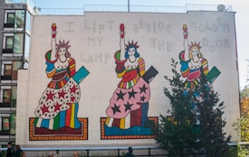 A large wall mural depicting three stylized figures resembling the Statue of Liberty in colorful, patterned dresses. Each figure is holding a torch and the mural features the phrase 'I lift my lamp beside the golden door.' The background is a plain, light-colored wall, and there are trees and people in the foreground.