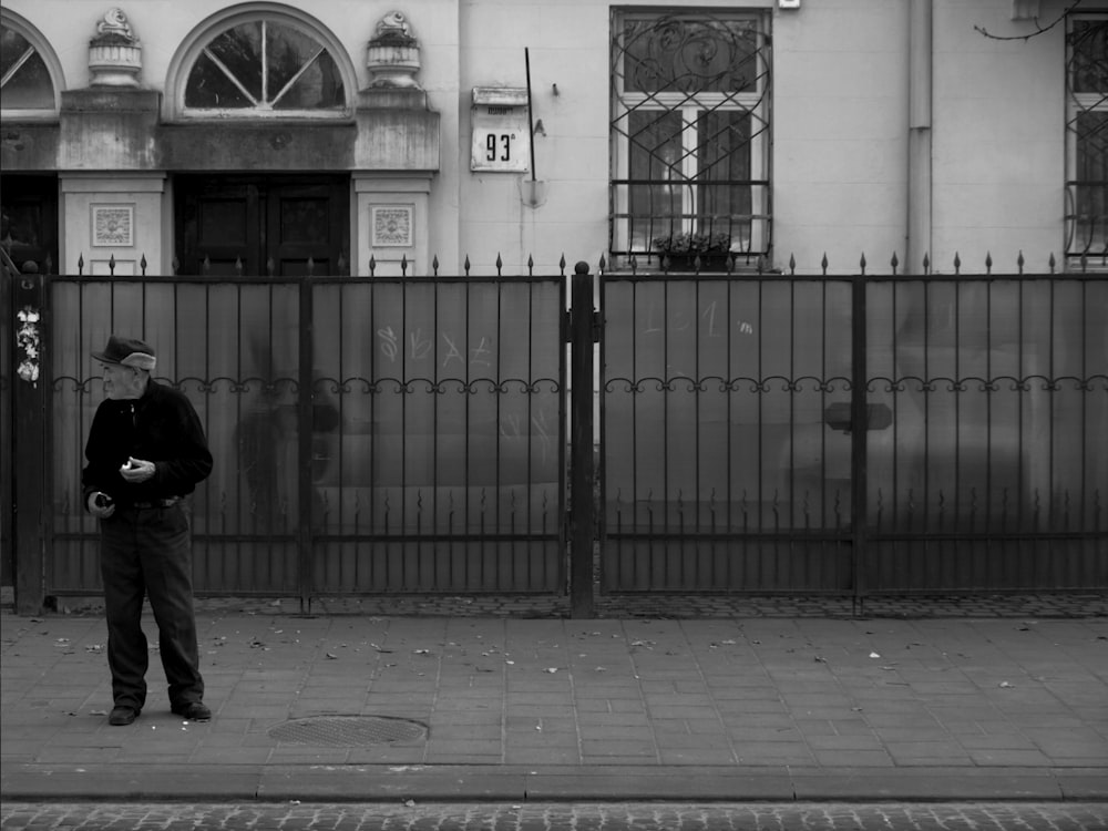 grayscale photo of man in black jacket standing beside metal gate