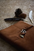 A brown leather journal with a metallic clover-shaped clasp and key rests on a textured surface. Two pine cones and two feathers, one white and one dark-colored, are placed near the journal.