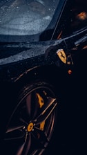 A close-up of a luxury sports car with visible raindrops on its sleek black surface. The focus is on the car's wheel and the iconic logo on the side, with ambient lighting highlighting the contours.