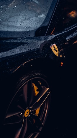 A close-up of a luxury sports car with visible raindrops on its sleek black surface. The focus is on the car's wheel and the iconic logo on the side, with ambient lighting highlighting the contours.