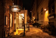 A dimly lit outdoor scene captures a narrow alleyway lined with brick buildings. On the left, a restaurant sign reads 'Restaurant & Bar' with an illuminated entrance porch. Warm lighting casts a soft glow on the facades and highlights greenery in planters along the path.
