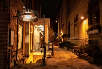 A dimly lit outdoor scene captures a narrow alleyway lined with brick buildings. On the left, a restaurant sign reads 'Restaurant & Bar' with an illuminated entrance porch. Warm lighting casts a soft glow on the facades and highlights greenery in planters along the path.