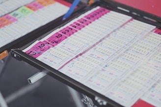 A collection of lottery tickets is organized neatly in a metal tray. The tickets display various numbers and have color-coded sections, primarily in pink and white. Some tickets have vibrant green sections, adding contrast to the overall layout.