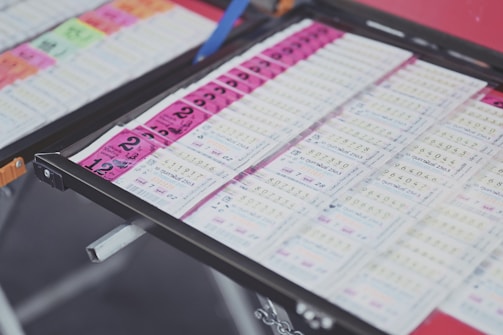 A collection of lottery tickets is organized neatly in a metal tray. The tickets display various numbers and have color-coded sections, primarily in pink and white. Some tickets have vibrant green sections, adding contrast to the overall layout.
