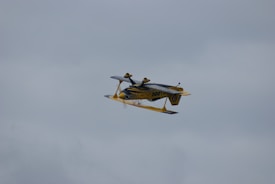A yellow and black aerobatic airplane is flying upside down against a backdrop of overcast sky. The aircraft's distinctive colors and design are visible as it performs the maneuver.