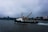 Several ships are visible on a body of water under an overcast sky. A prominent yellow crane is mounted on a black and white vessel in the foreground. Other ships, including a warship, are anchored in the background near industrial structures along the shoreline.