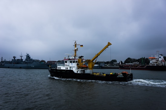Several ships are visible on a body of water under an overcast sky. A prominent yellow crane is mounted on a black and white vessel in the foreground. Other ships, including a warship, are anchored in the background near industrial structures along the shoreline.