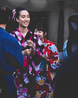Two women are wearing colorful traditional kimonos, adorned with floral patterns, standing alongside other people. One wears a black kimono with pink and white designs, and hair adorned with pink accessories, while the other wears a vibrant red kimono with yellow and blue patterns. The scene suggests a cultural or festive event.