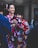 Two women are wearing colorful traditional kimonos, adorned with floral patterns, standing alongside other people. One wears a black kimono with pink and white designs, and hair adorned with pink accessories, while the other wears a vibrant red kimono with yellow and blue patterns. The scene suggests a cultural or festive event.