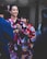 Two women are wearing colorful traditional kimonos, adorned with floral patterns, standing alongside other people. One wears a black kimono with pink and white designs, and hair adorned with pink accessories, while the other wears a vibrant red kimono with yellow and blue patterns. The scene suggests a cultural or festive event.