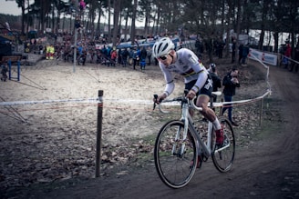 A cyclist wearing a white racing outfit and helmet is intensely riding a bicycle on a dirt path through a wooded area. The path is surrounded by spectators, some of whom are taking photos. Fallen leaves cover the ground, and a barrier runs alongside the path.