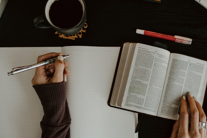 Close-up of hands writing in a notebook with an open Bible, coffee, and red pen, reflecting growth