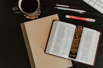 An open book with text revealing pages from the Bible is placed on a dark table. A decorative bookmark with religious text is positioned inside the book. A closed notebook lies underneath the book. Nearby, there are pens, a highlighter, a mug filled with a dark beverage, and part of a computer keyboard is visible in the top right corner.