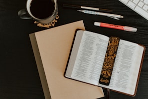 An open book with text revealing pages from the Bible is placed on a dark table. A decorative bookmark with religious text is positioned inside the book. A closed notebook lies underneath the book. Nearby, there are pens, a highlighter, a mug filled with a dark beverage, and part of a computer keyboard is visible in the top right corner.