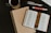 An open book with text revealing pages from the Bible is placed on a dark table. A decorative bookmark with religious text is positioned inside the book. A closed notebook lies underneath the book. Nearby, there are pens, a highlighter, a mug filled with a dark beverage, and part of a computer keyboard is visible in the top right corner.
