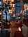 A restaurant interior warmly lit by clusters of decorative hanging lanterns. The space features large windows with a view of a building outside. Cozy seating includes red sofas and neutral-colored chairs, with wooden floors adding to the ambiance. There's a mix of modern and rustic elements, with natural touches like potted plants.