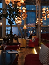 A restaurant interior warmly lit by clusters of decorative hanging lanterns. The space features large windows with a view of a building outside. Cozy seating includes red sofas and neutral-colored chairs, with wooden floors adding to the ambiance. There's a mix of modern and rustic elements, with natural touches like potted plants.