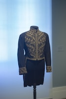 A black, intricately embroidered waistcoat is displayed on a mannequin against a light blue background. The detailed gold embroidery features ornate leaf patterns on the chest, collar, and cuffs.