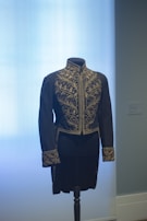 A black, intricately embroidered waistcoat is displayed on a mannequin against a light blue background. The detailed gold embroidery features ornate leaf patterns on the chest, collar, and cuffs.