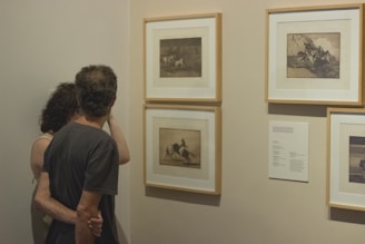 Two people are closely observing framed artwork displayed on a wall. The artwork consists of four framed pieces, possibly sketches or prints with subjects that appear to have a classical or historical theme. The couple is standing with their backs to the viewer, with one arm around the other, indicating a shared interest in the art.