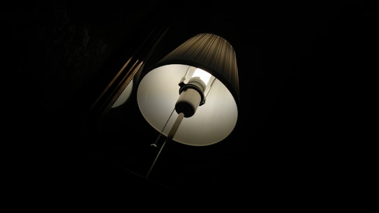 A lampshade illuminated in a dark environment, casting a warm glow. The minimal light emphasizes the pleated texture of the shade, creating contrast with the surrounding darkness.