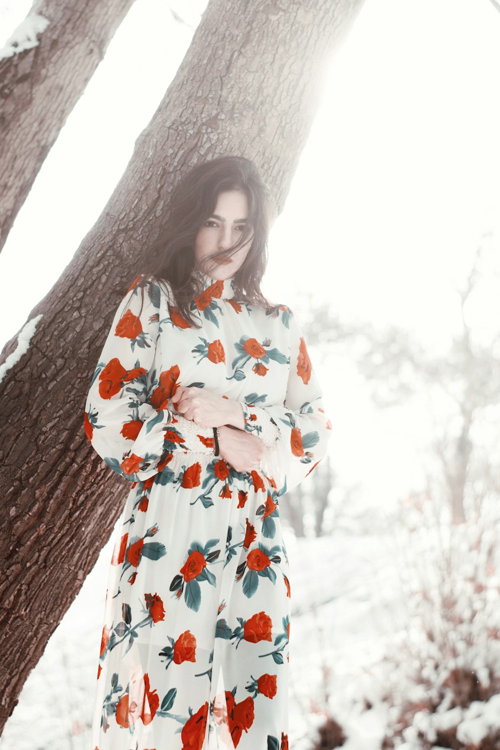 Femme en robe fleurie blanche, rouge et verte debout à côté d’un arbre brun