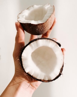A hand is holding two halves of a coconut. The interior is white and the outer shell is brown. The light casts soft shadows, highlighting the fibrous texture of the shell.