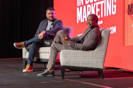 Two individuals are seated on stage in upholstered chairs, engaged in a discussion. The background features a bright red display with text related to marketing. Both are dressed in business casual attire.