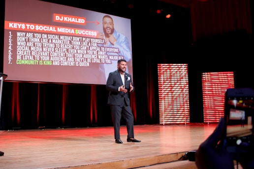 A man in a suit is speaking on stage with a large presentation slide behind him titled 'Keys to Social Media Success'. The slide lists several bullet points on successful social media strategies. The stage is lit with red and orange tones, and another part of the stage features striped panels emitting red light.