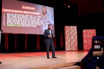 A man in a suit is speaking on stage with a large presentation slide behind him titled 'Keys to Social Media Success'. The slide lists several bullet points on successful social media strategies. The stage is lit with red and orange tones, and another part of the stage features striped panels emitting red light.