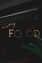 A neon sign with the words 'healthy FOOD' is illuminated against a dark background. The sign is partially obscured by an awning above it. In the foreground, part of a green plant is visible.