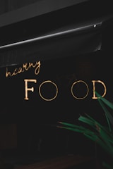 A neon sign with the words 'healthy FOOD' is illuminated against a dark background. The sign is partially obscured by an awning above it. In the foreground, part of a green plant is visible.