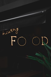 A neon sign with the words 'healthy FOOD' is illuminated against a dark background. The sign is partially obscured by an awning above it. In the foreground, part of a green plant is visible.