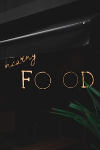 A neon sign with the words 'healthy FOOD' is illuminated against a dark background. The sign is partially obscured by an awning above it. In the foreground, part of a green plant is visible.