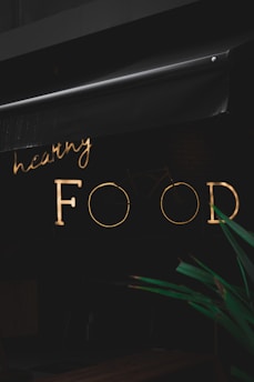 A neon sign with the words 'healthy FOOD' is illuminated against a dark background. The sign is partially obscured by an awning above it. In the foreground, part of a green plant is visible.