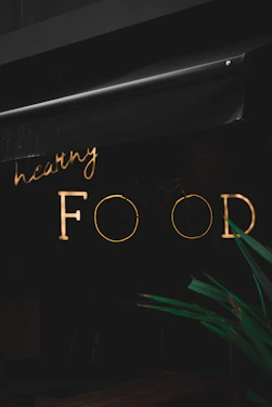 A neon sign with the words 'healthy FOOD' is illuminated against a dark background. The sign is partially obscured by an awning above it. In the foreground, part of a green plant is visible.