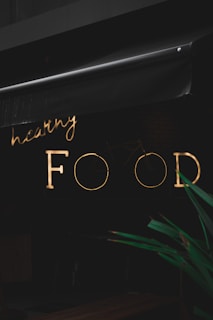 A neon sign with the words 'healthy FOOD' is illuminated against a dark background. The sign is partially obscured by an awning above it. In the foreground, part of a green plant is visible.