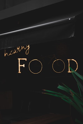 A neon sign with the words 'healthy FOOD' is illuminated against a dark background. The sign is partially obscured by an awning above it. In the foreground, part of a green plant is visible.