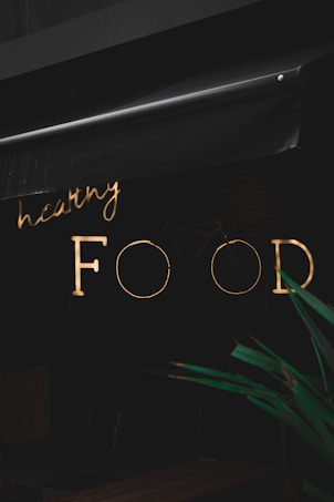A neon sign with the words 'healthy FOOD' is illuminated against a dark background. The sign is partially obscured by an awning above it. In the foreground, part of a green plant is visible.