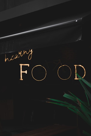 A neon sign with the words 'healthy FOOD' is illuminated against a dark background. The sign is partially obscured by an awning above it. In the foreground, part of a green plant is visible.