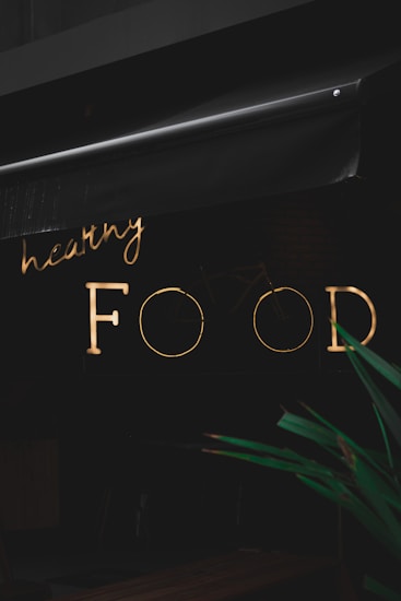A neon sign with the words 'healthy FOOD' is illuminated against a dark background. The sign is partially obscured by an awning above it. In the foreground, part of a green plant is visible.
