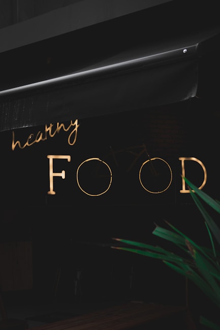 A neon sign with the words 'healthy FOOD' is illuminated against a dark background. The sign is partially obscured by an awning above it. In the foreground, part of a green plant is visible.