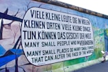A graffiti mural on a large wall with a message written in German and English. The German text reads, 'Viele kleine Leute die in vielen kleinen Orten viele kleine Dinge tun, können das Gesicht der Welt,' which translates to the English text below it, 'Many small people who in many small places do many small things that can alter the face of the world.' The mural features a colorful background with abstract images of trees and landscapes.